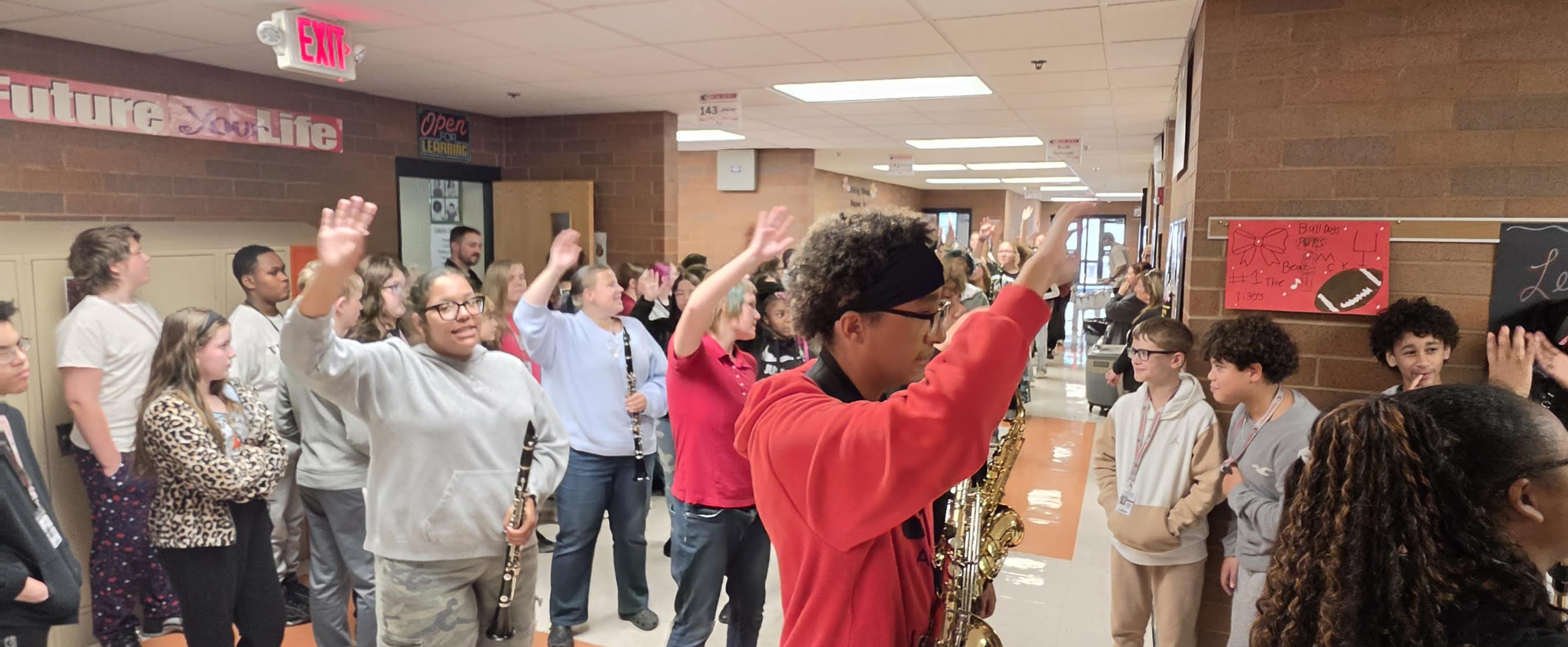 A group of students stands in a school hallway, with many raising their hands or waving while holding musical instruments.