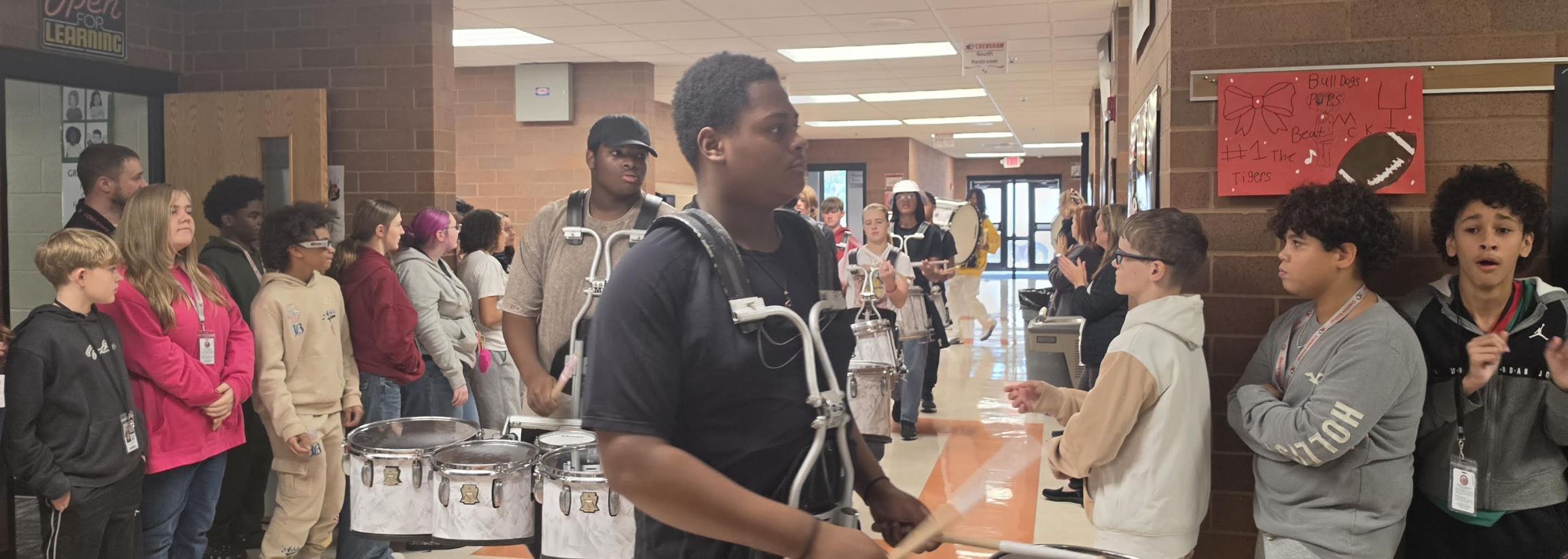A school hallway filled with students, some wearing marching band drums and harnesses.
