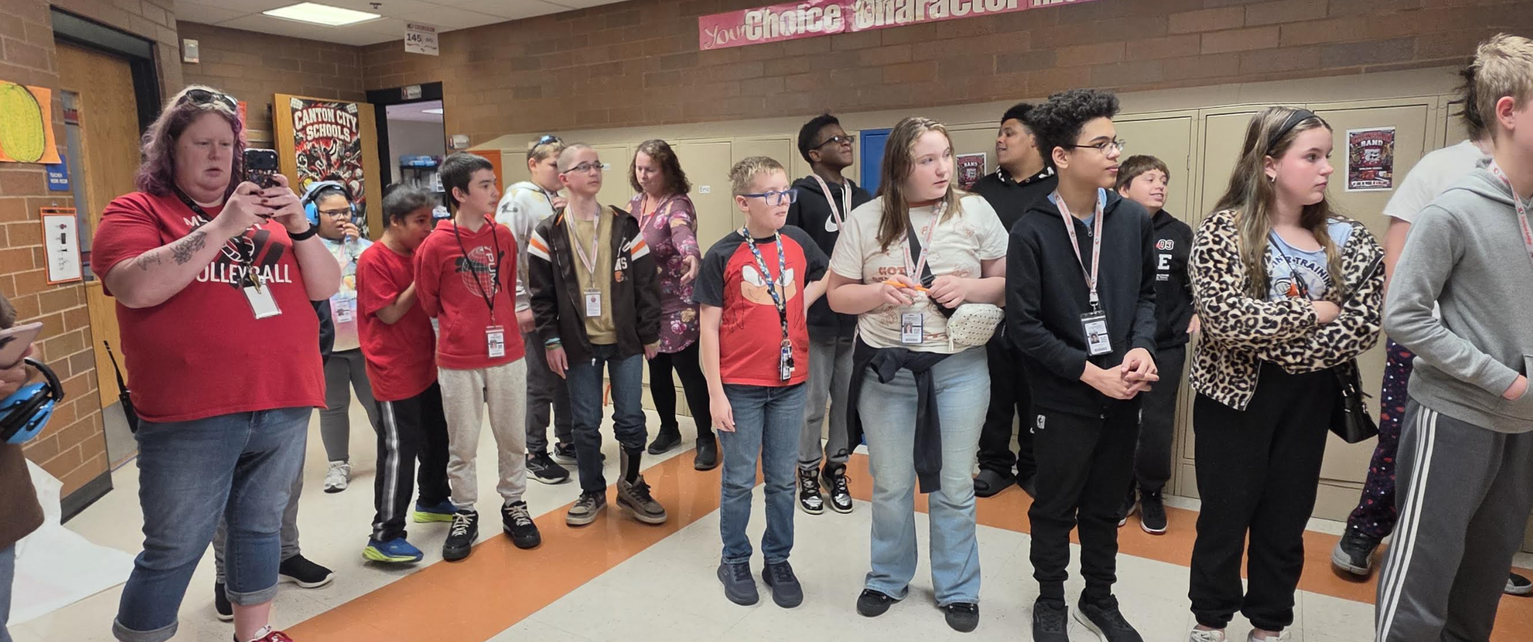 A group of students and teachers stand in a school hallway near lockers, looking towards the right side of the frame.