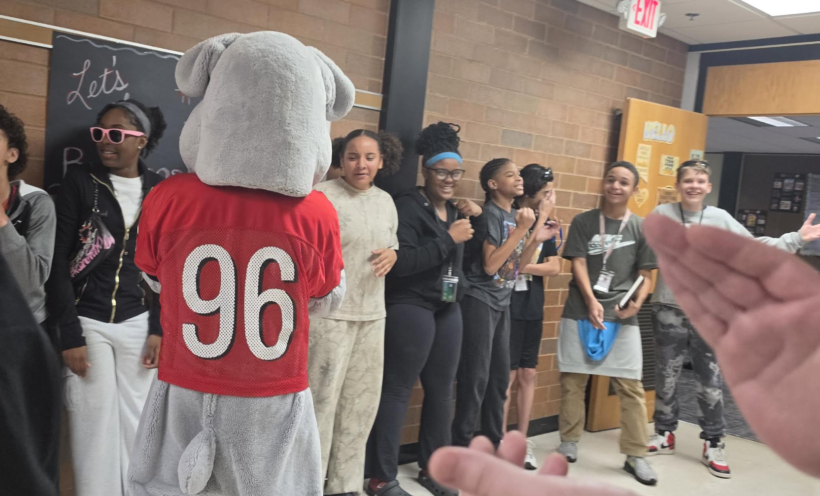 A school mascot in a red jersey with the number 96 stands facing a line of smiling students in a hallway.