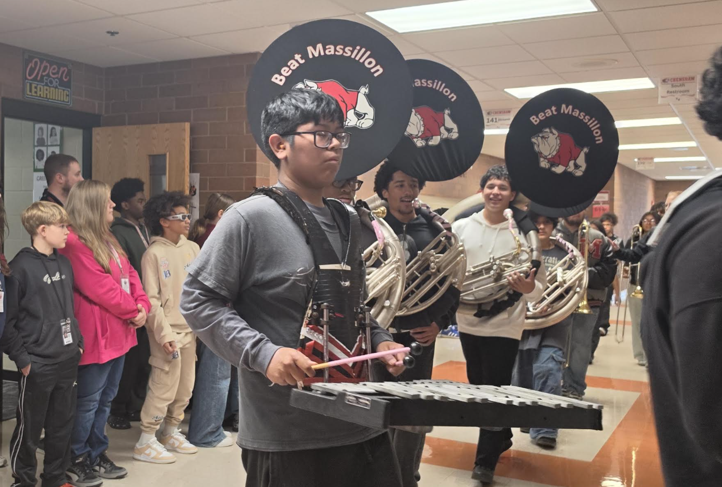 A high school marching band walks through a hallway, with a student in the foreground carrying a glockenspiel.