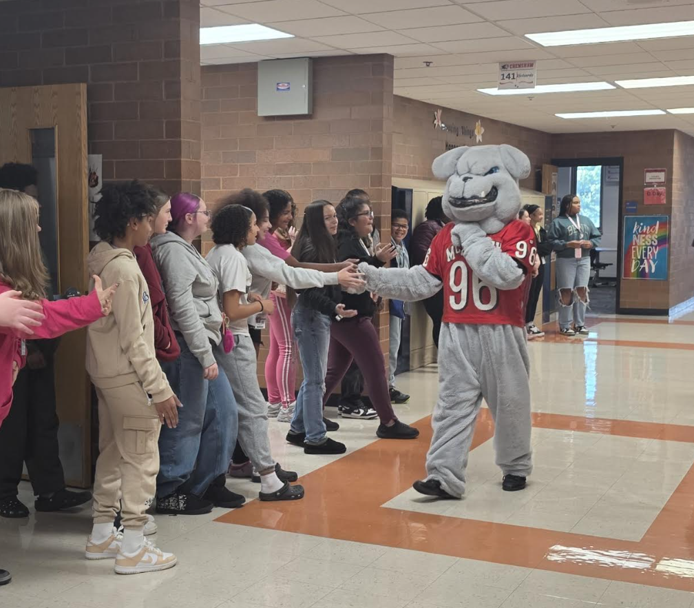 A school mascot dressed as a grey bulldog in a red jersey high-fives students in a hallway.