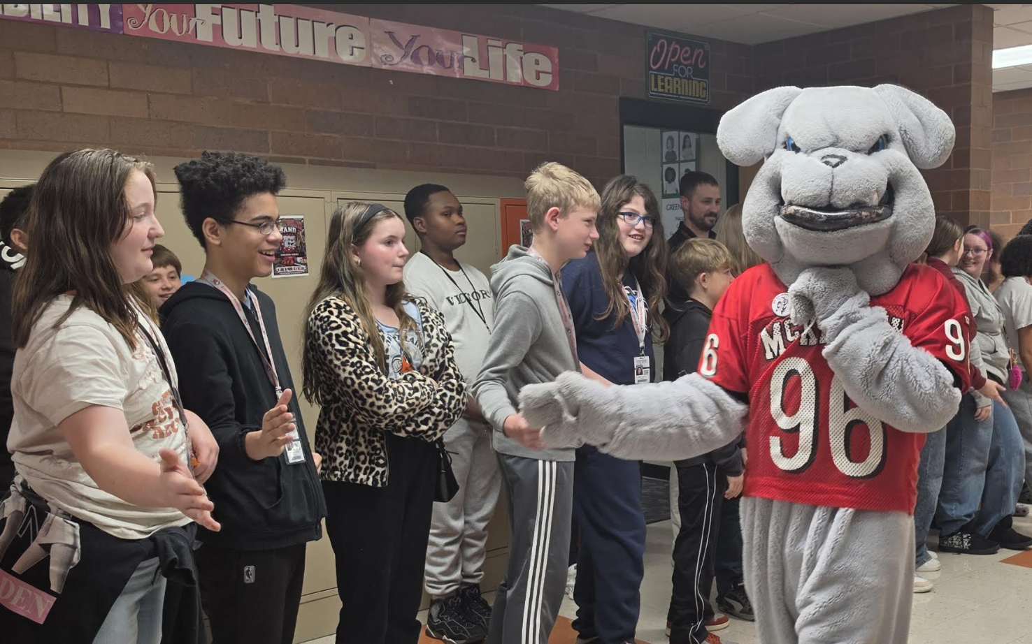 A bulldog mascot wearing a red jersey with the number 96 stands in a school hallway greeting students.