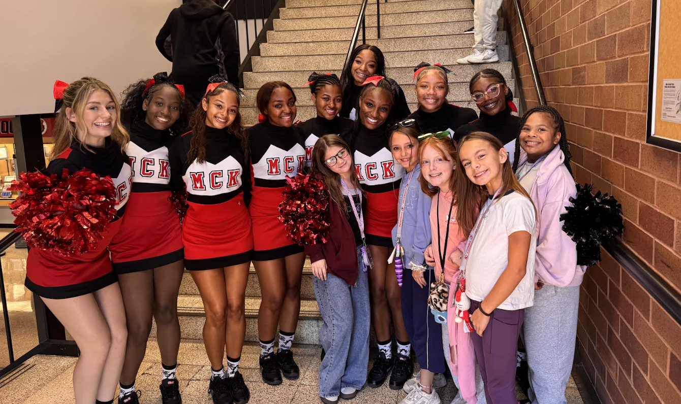 A group of cheerleaders and students in red, white, and black uniforms stand on a staircase, smiling.