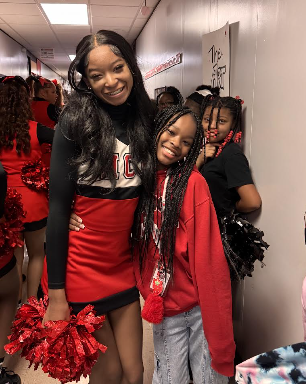 Two smiling girls pose together in a school hallway, with one in a red and black cheerleader uniform holding pom-poms.