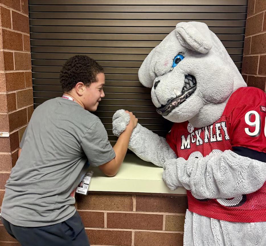 A smiling young person arm wrestles with a large grey bulldog mascot wearing a red jersey with the text "MCKINLEY 90".