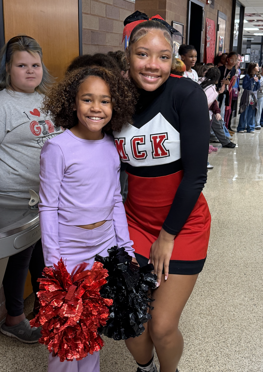A young girl in a purple outfit holds red and black pompoms next to a smiling cheerleader in a red, black, and white uniform inside a school hallway.