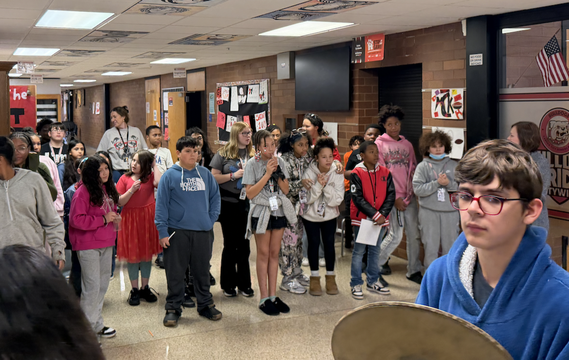 A group of students stand attentively in a brightly lit school hallway.