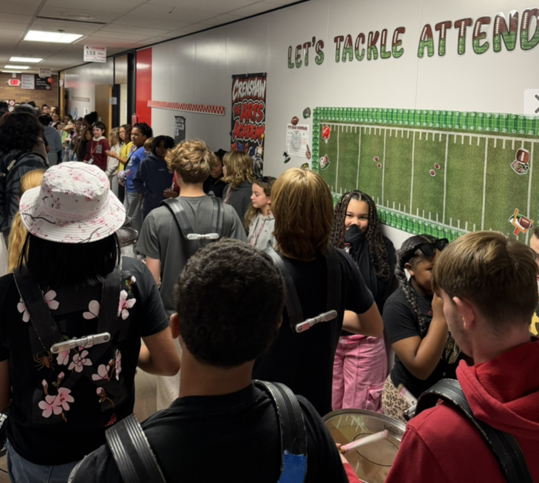 A line of students with marching band instruments stands in a school hallway with football-themed decor.