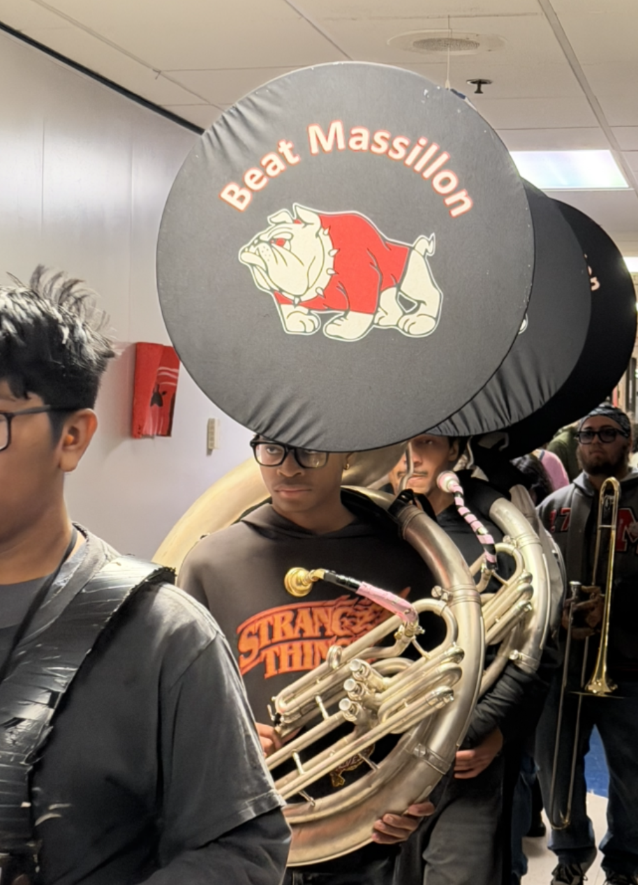 A line of marching band members carries large sousaphones down an indoor hallway, with the front instrument featuring a bulldog graphic and the text "Beat Massillon."
