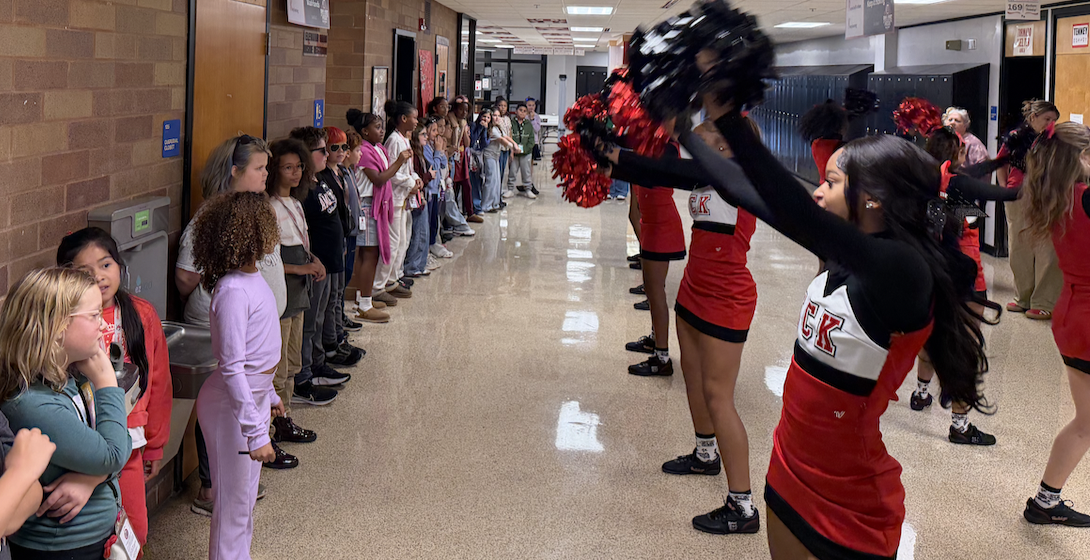 Cheerleaders in red and white uniforms perform with pom-poms in a crowded school hallway, while students watch.