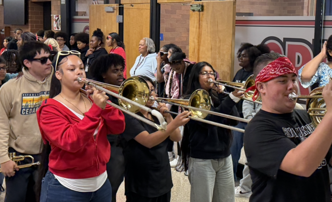 A diverse group of students in a hallway play trombones, focused on their music.