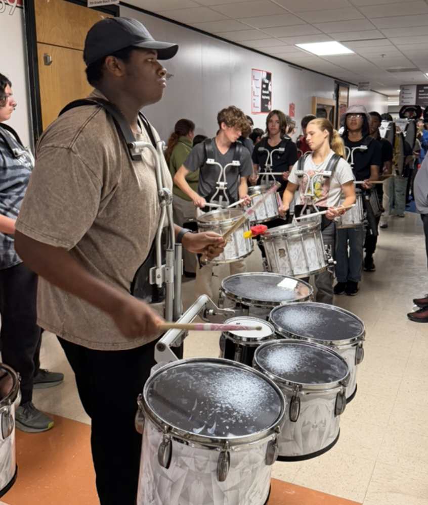 A marching band percussion section lines up in a school hallway, wearing harness drums and holding drumsticks.
