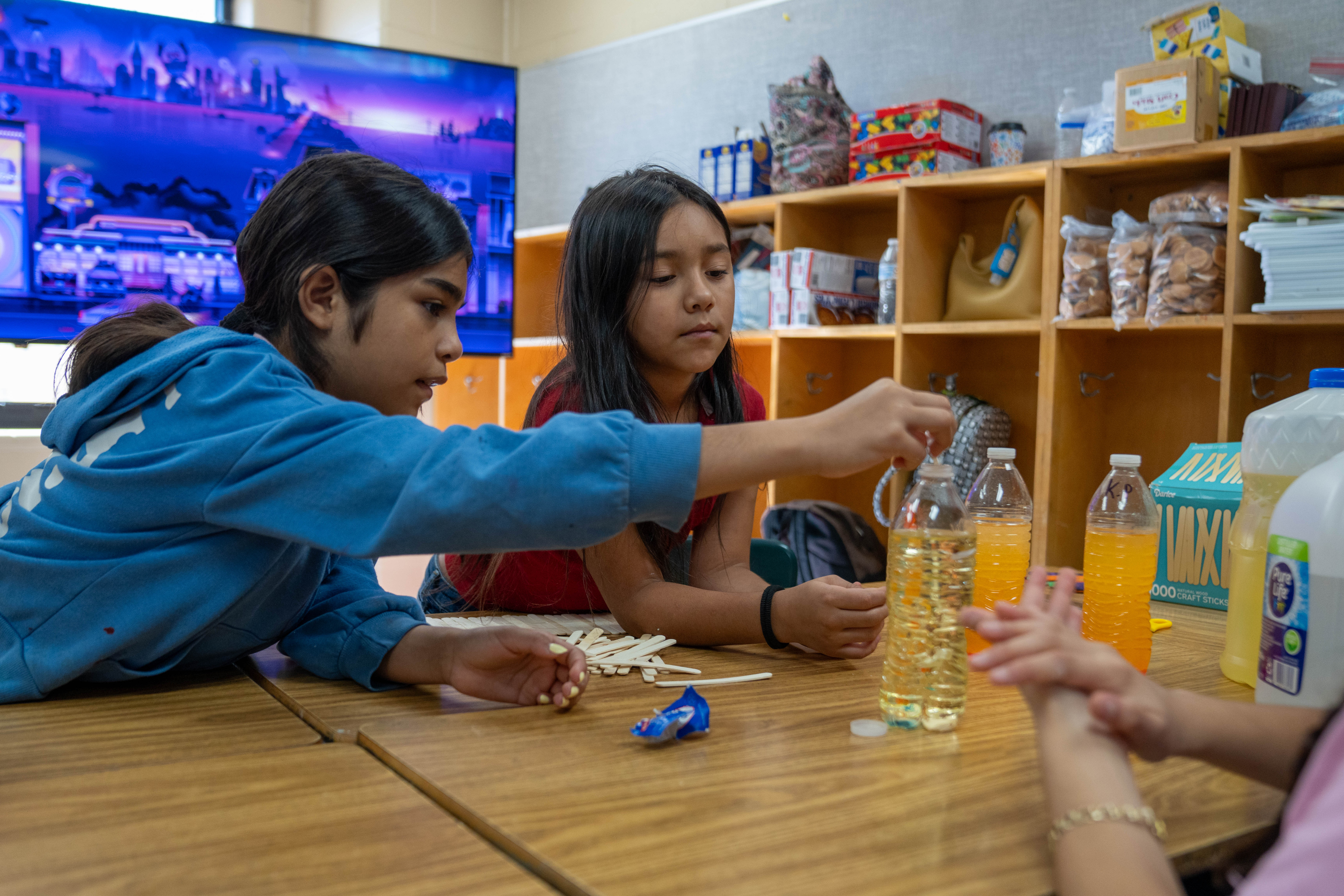 Two children are engaged in a hands-on activity at a wooden table, with bottles of liquid and craft sticks in front of them.