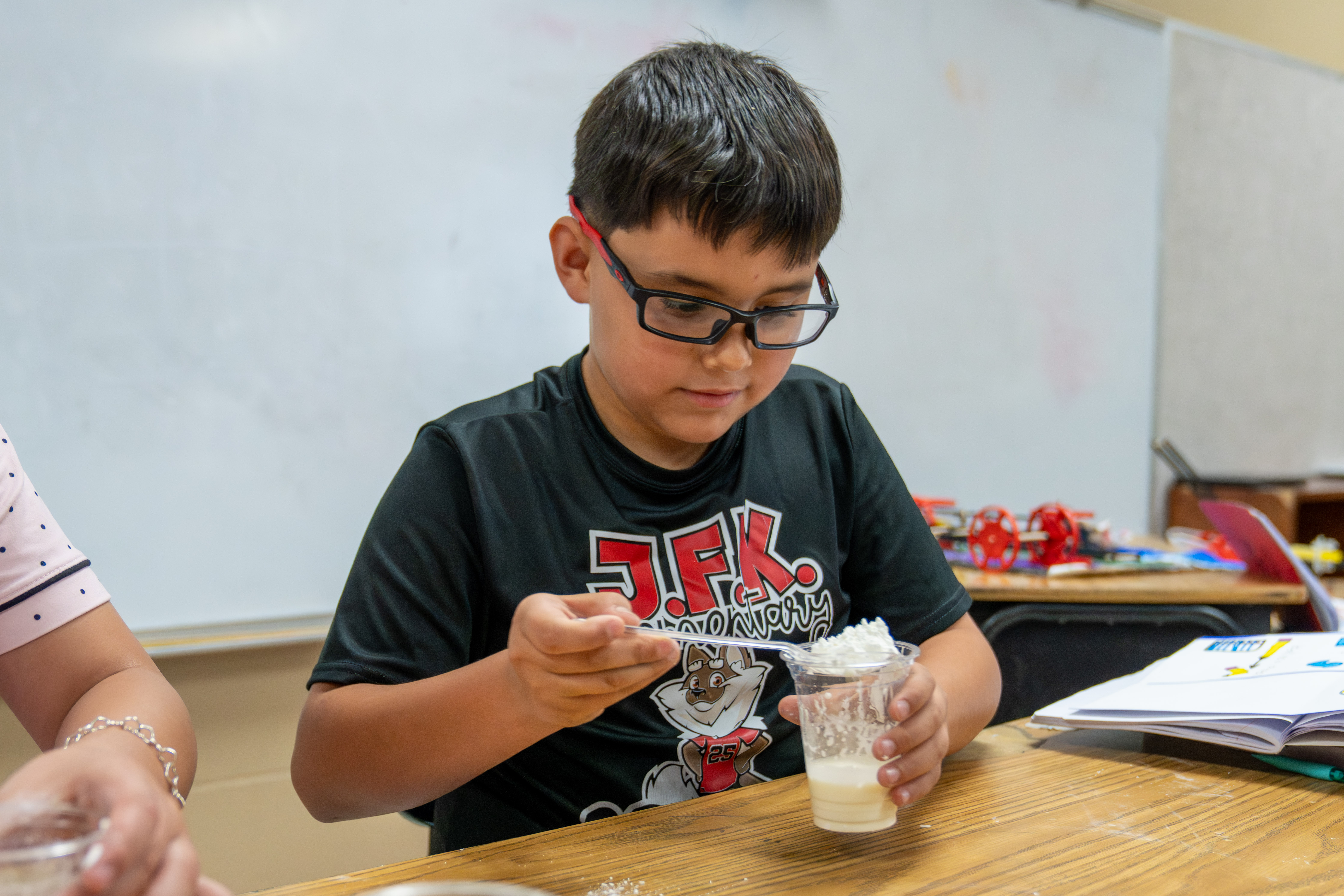 A young boy wearing glasses and a black t-shirt with a fox logo is adding whipped cream to a clear cup.