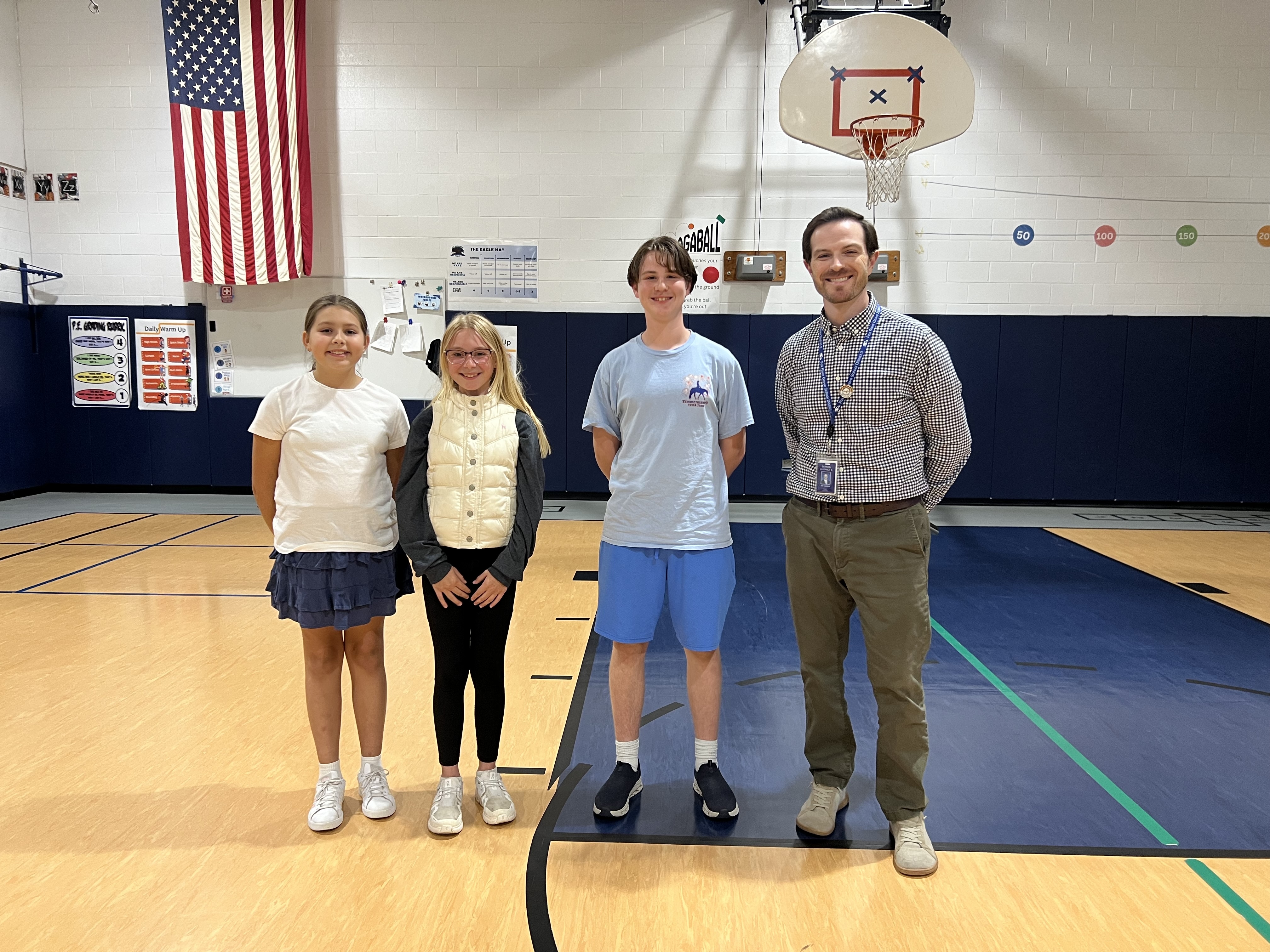 Three students and an adult teacher stand smiling inside a school gymnasium with an American flag on the wall.