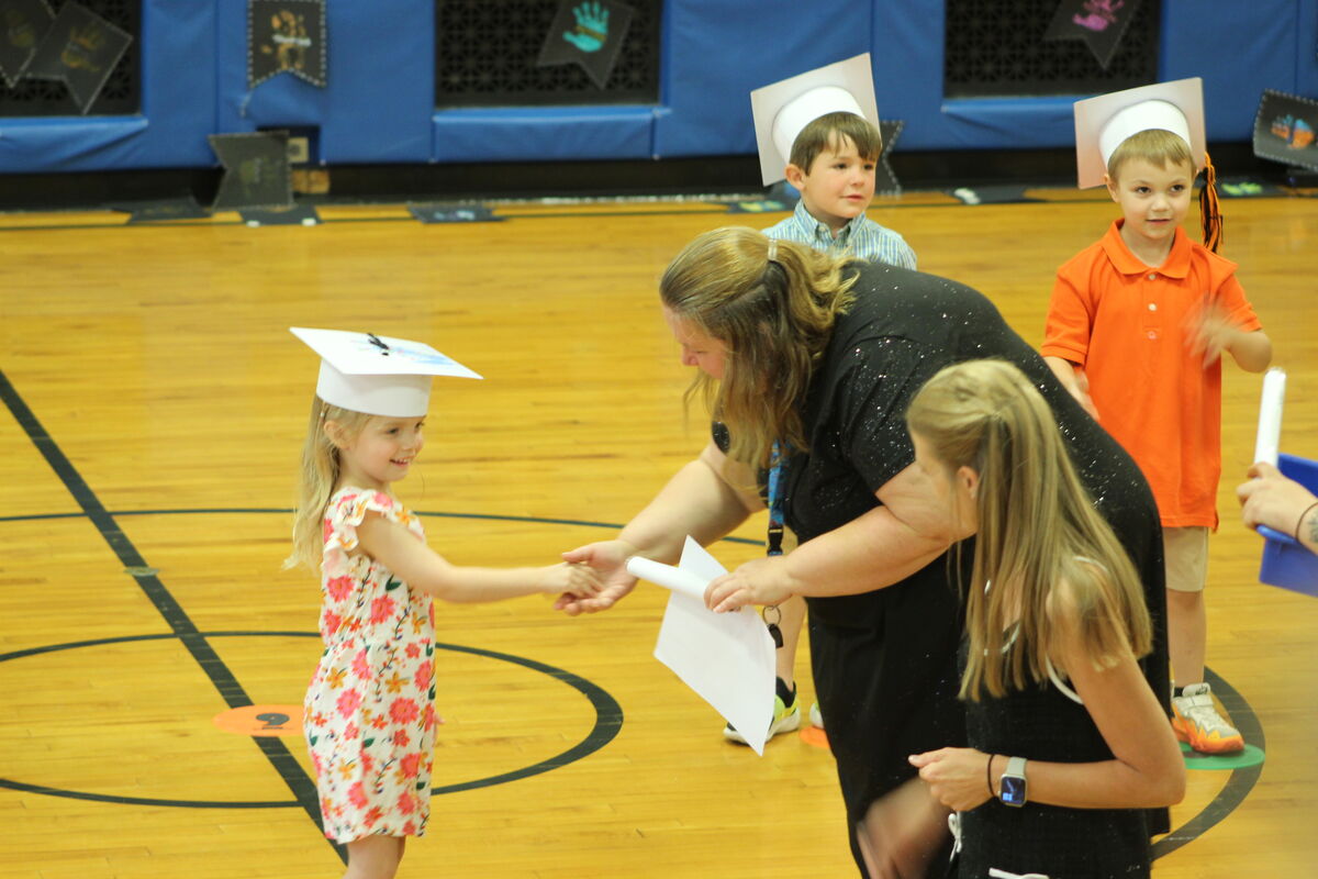 A teacher congratulates a young girl in a floral dress and graduation cap on a gymnasium floor.
