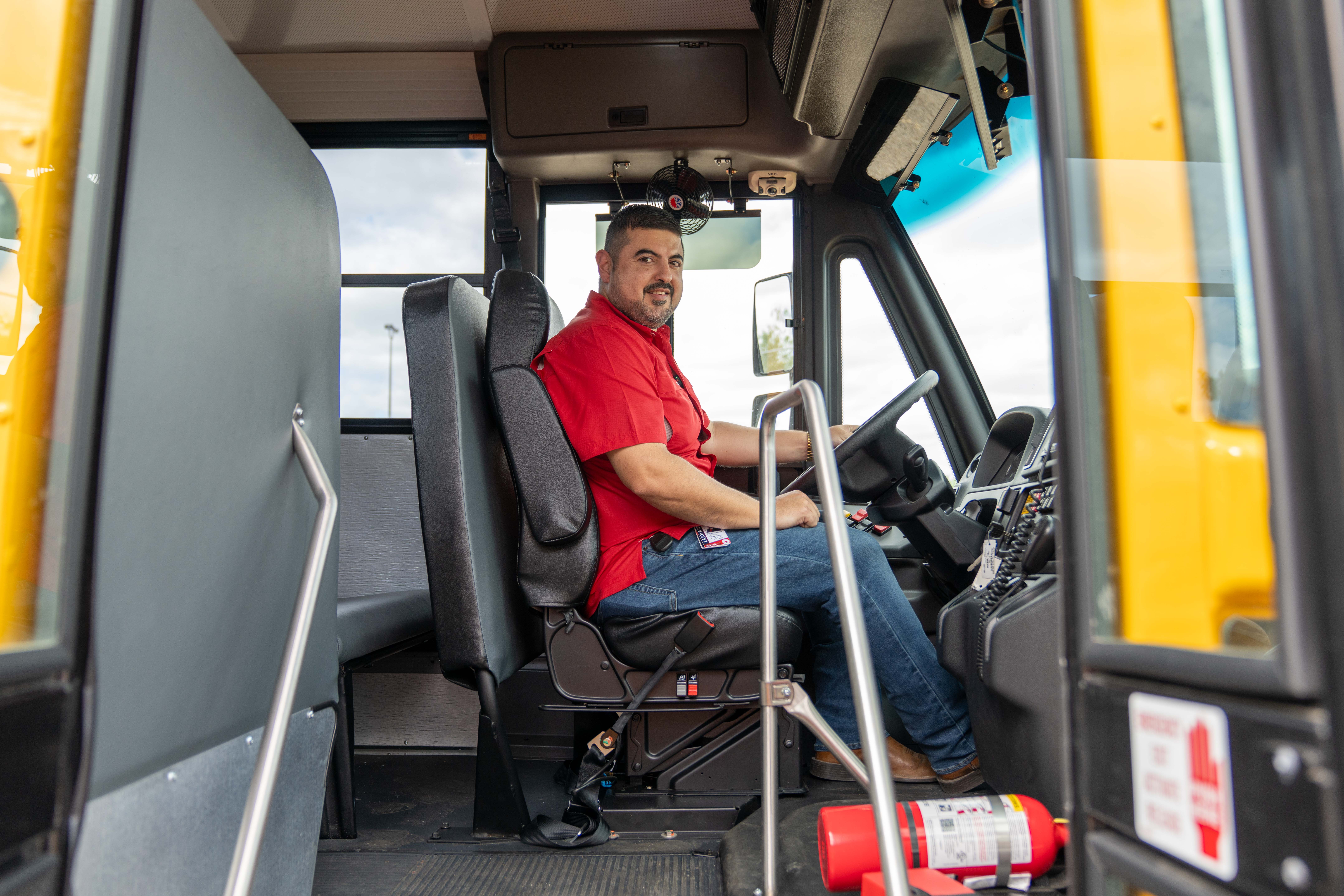 A friendly school bus driver in a red shirt sits in the driver's seat, smiling at the camera.