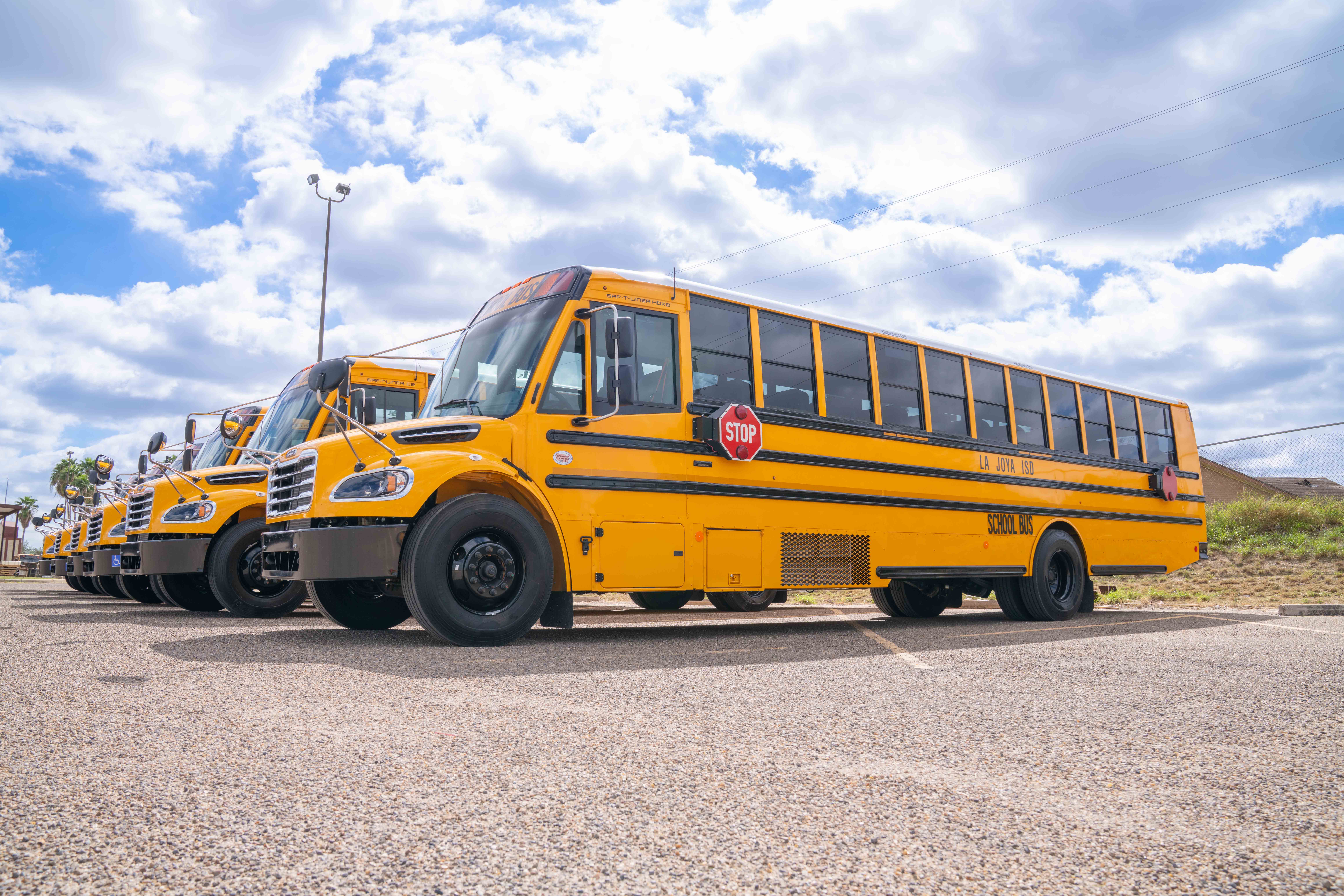 A row of bright yellow school buses is parked neatly in a gravel lot under a cloudy blue sky.