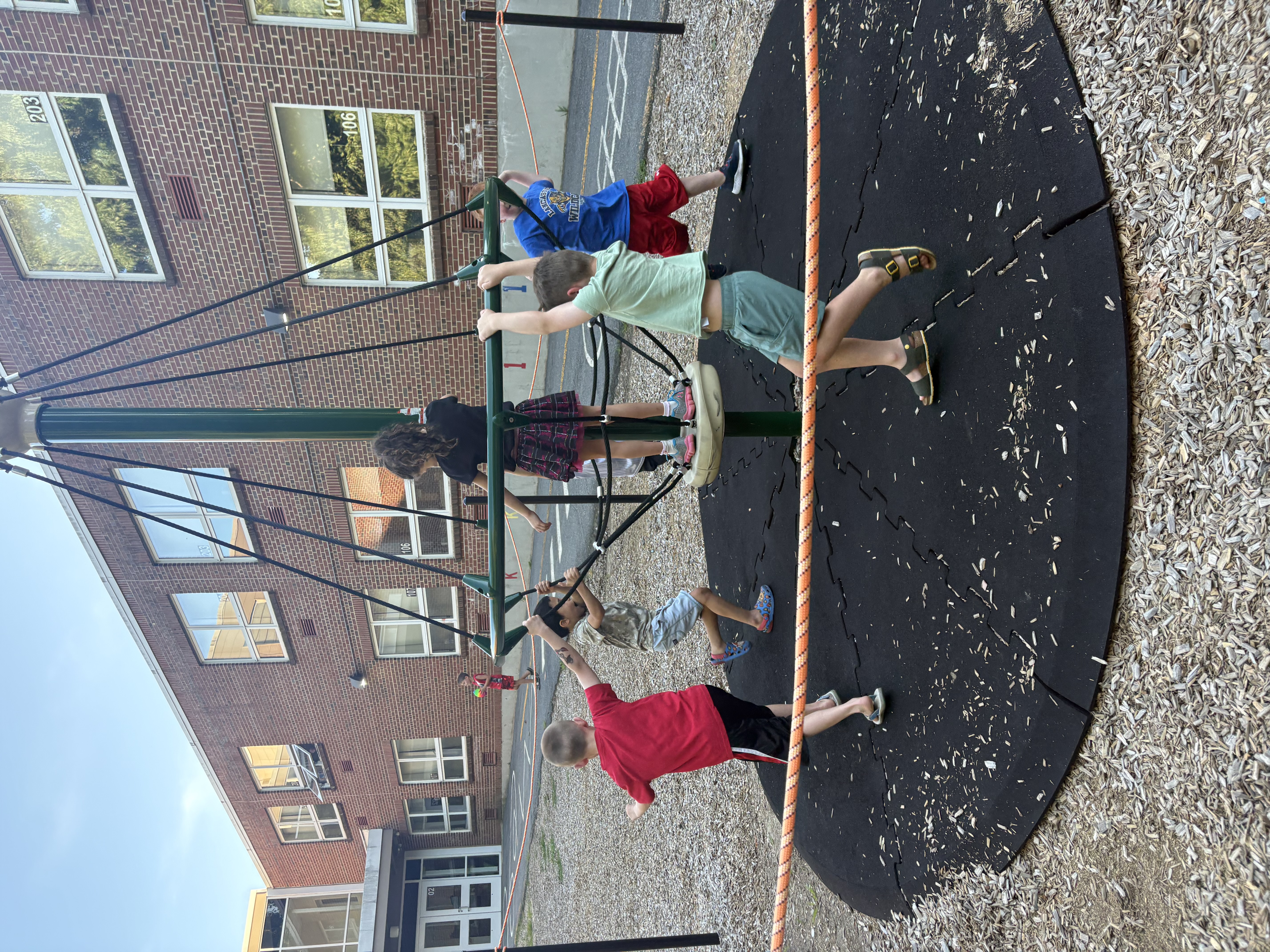 Children play on a merry-go-round on a playground with a brick building in the background.