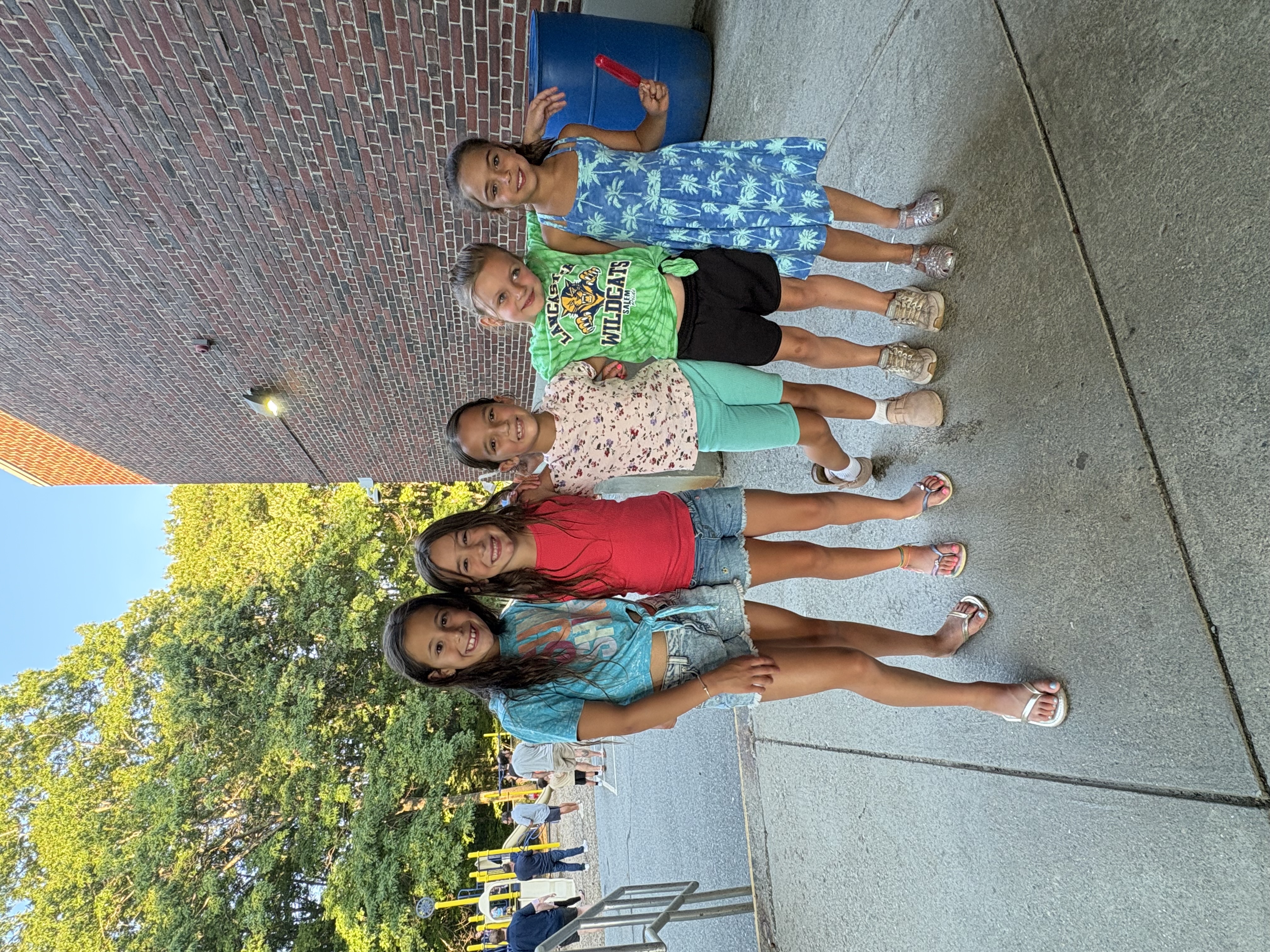 Four smiling girls stand together outdoors, with one holding a red popsicle.