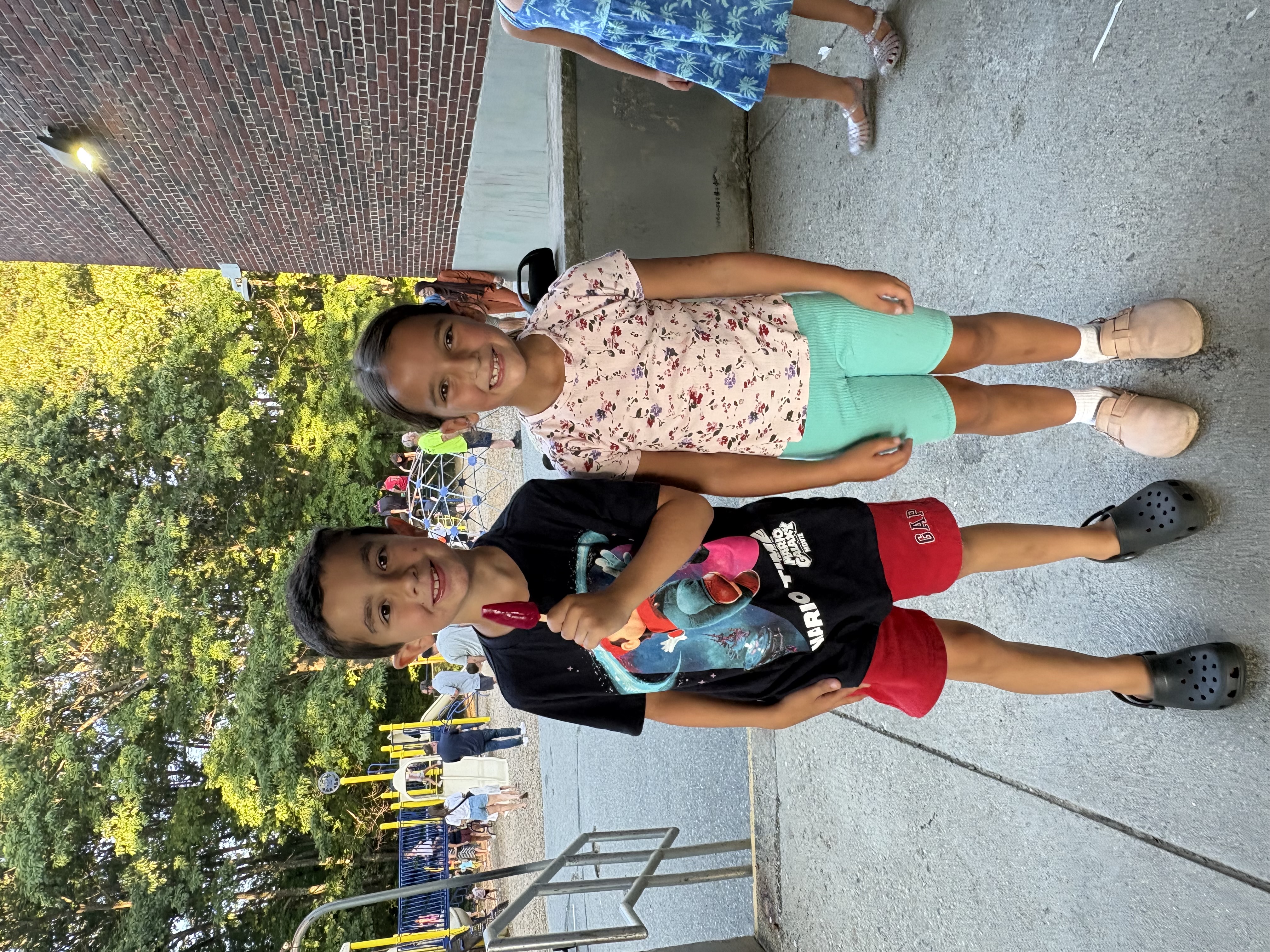 Two smiling children stand outdoors, one holding a red popsicle, with a playground in the background.