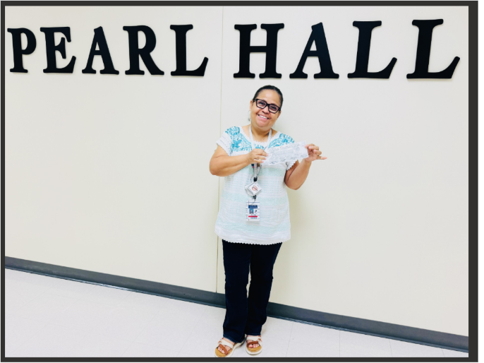 A smiling woman wearing glasses and a lanyard stands in front of a white wall with the text 'PEARL HALL', holding up a paper in both hands.