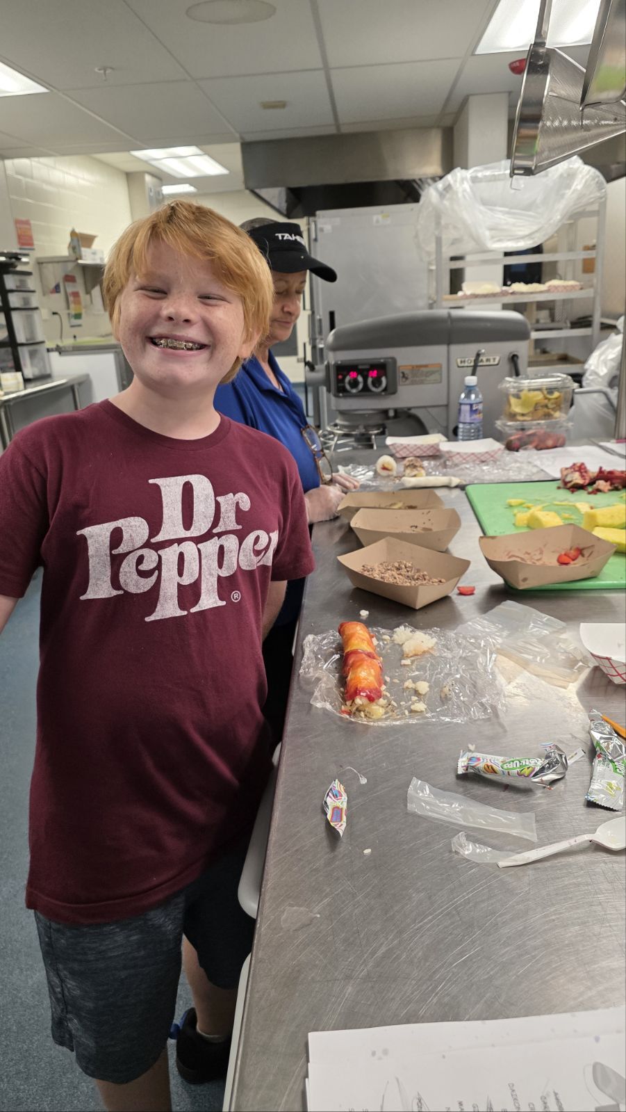 A smiling boy wearing a Dr Pepper shirt stands in a kitchen with food preparation stations.
