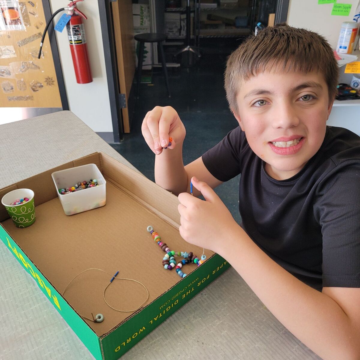 A young boy smiles while making jewelry with colorful beads at a table.