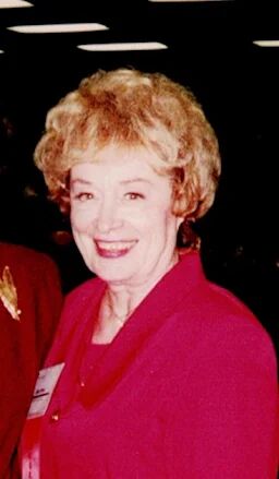A woman with blonde, curly hair smiles warmly, wearing a bright pink jacket.