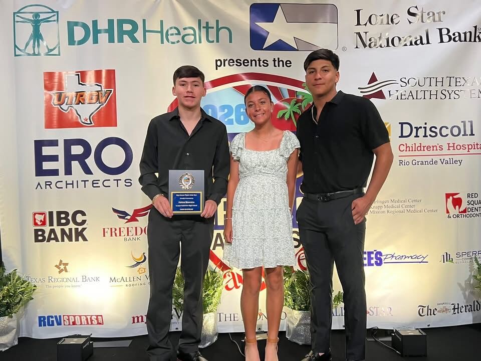 Three young people stand on a stage in front of a backdrop with sponsor logos, one holding an award.