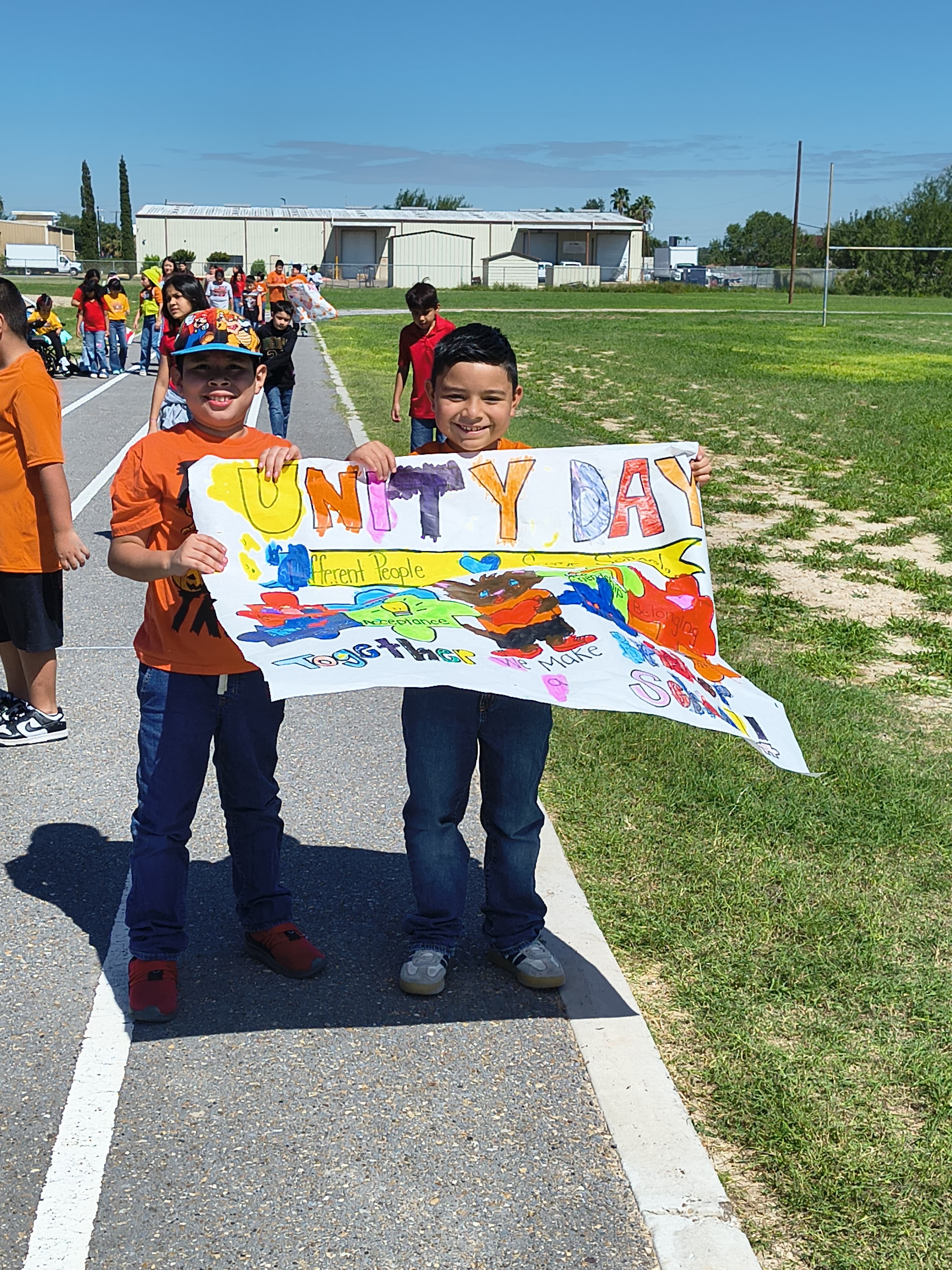 Two smiling students proudly hold a colorful hand-painted banner that reads "Unity Day" outdoors on a sunny day.