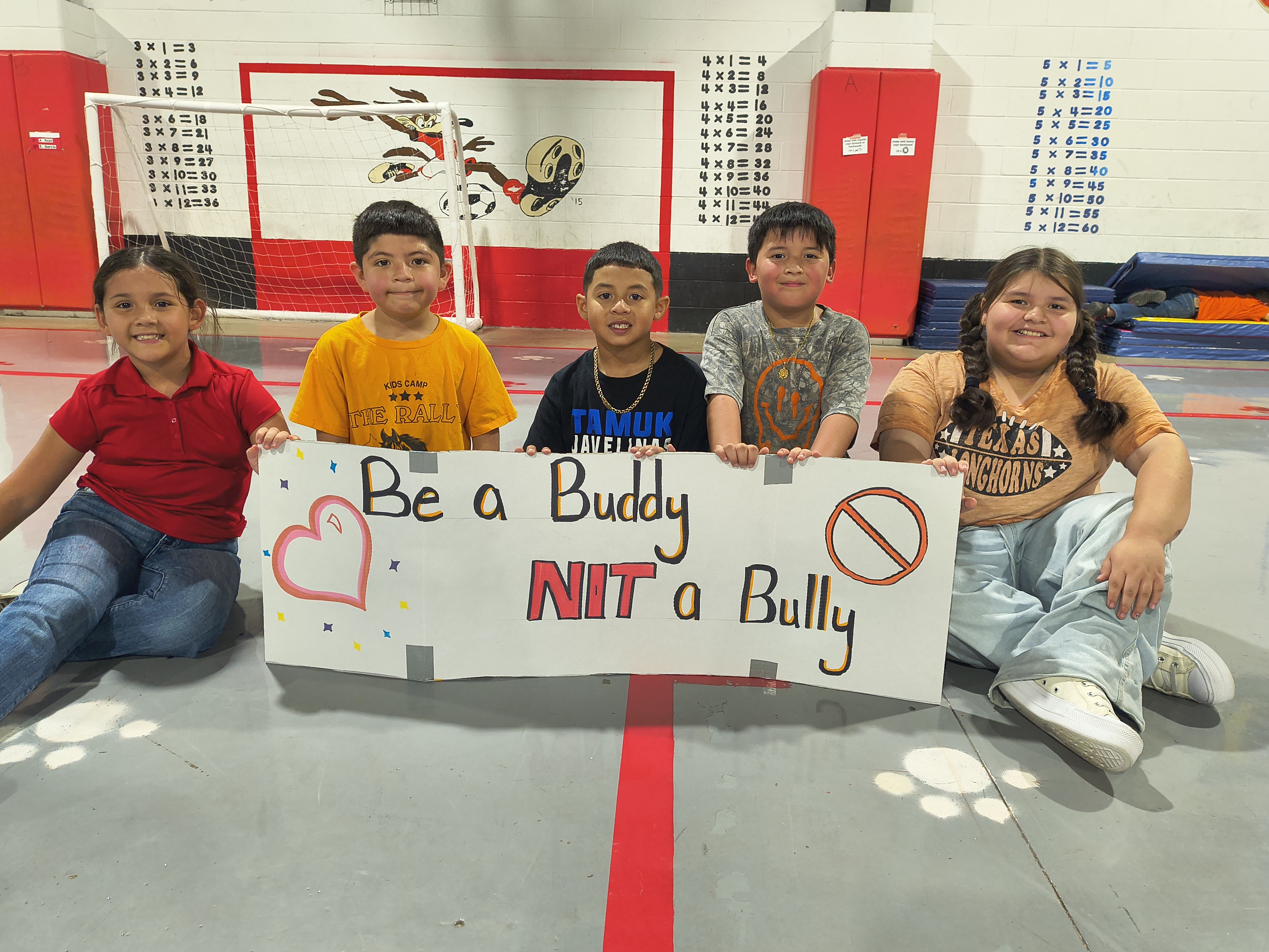 Five smiling students sit in a gym holding a homemade sign that reads, Be a Buddy NOT a Bully.