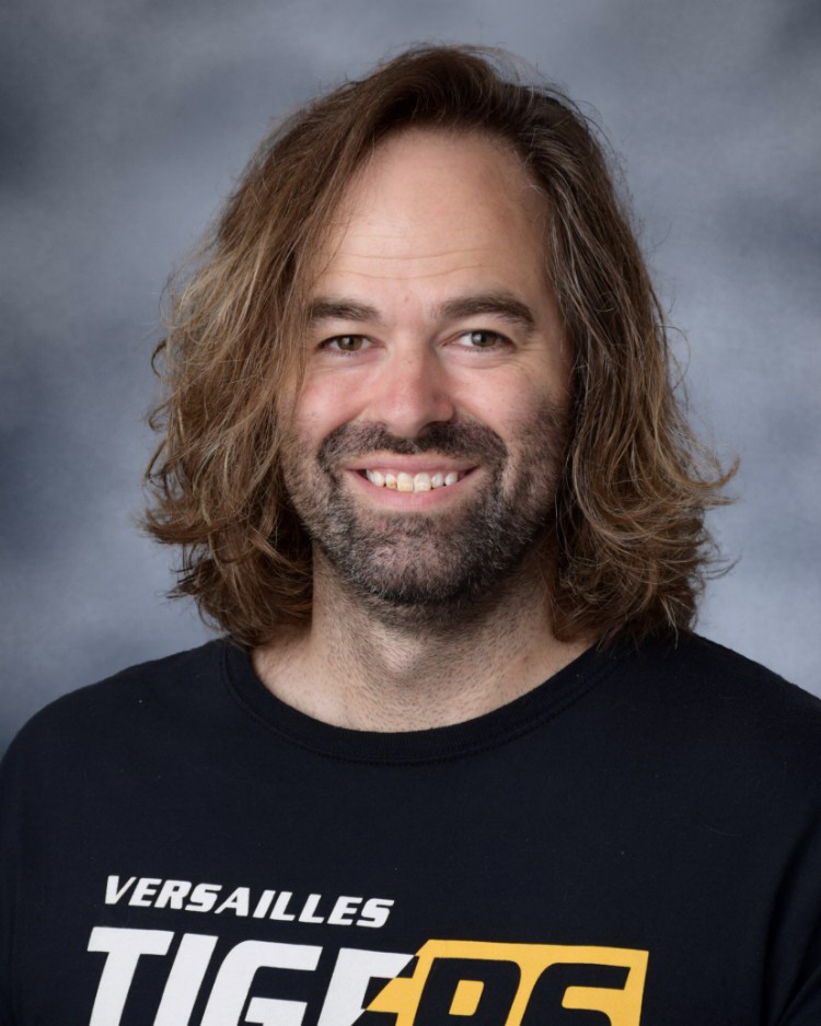 A smiling man with long brown hair and a beard wears a black t-shirt with the text 'VERSAILLES TIGERS'.