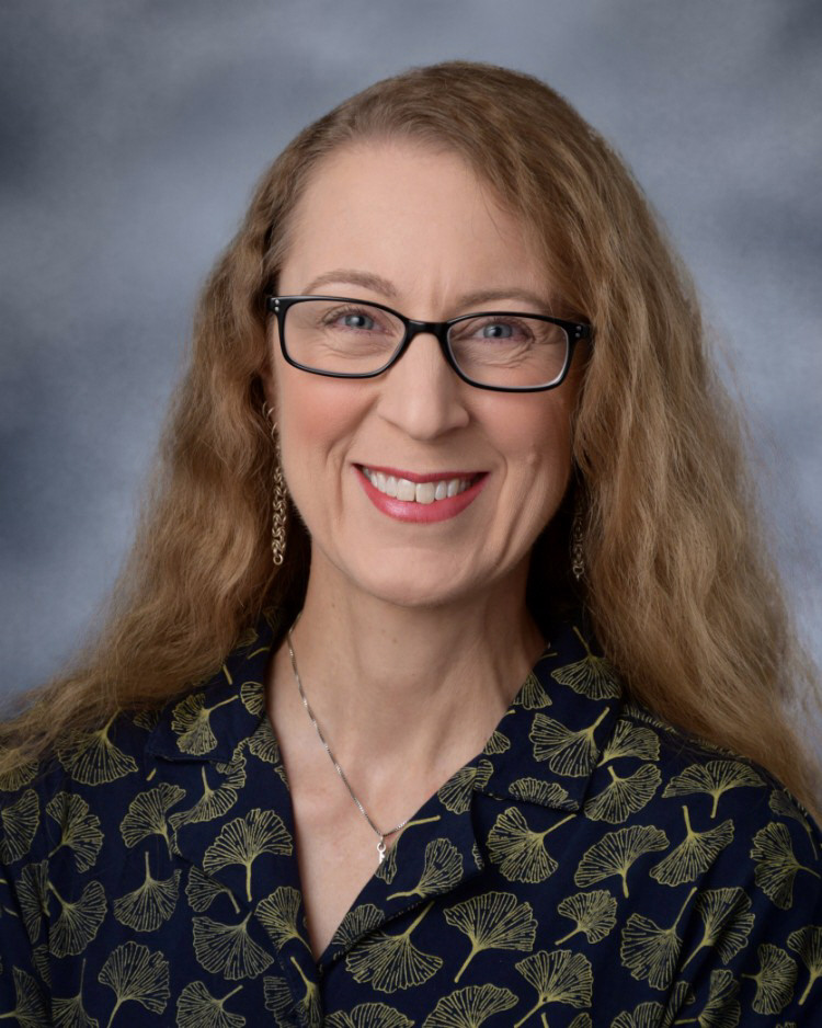 A professional headshot of a smiling woman with long blonde hair, wearing glasses and a patterned blouse.