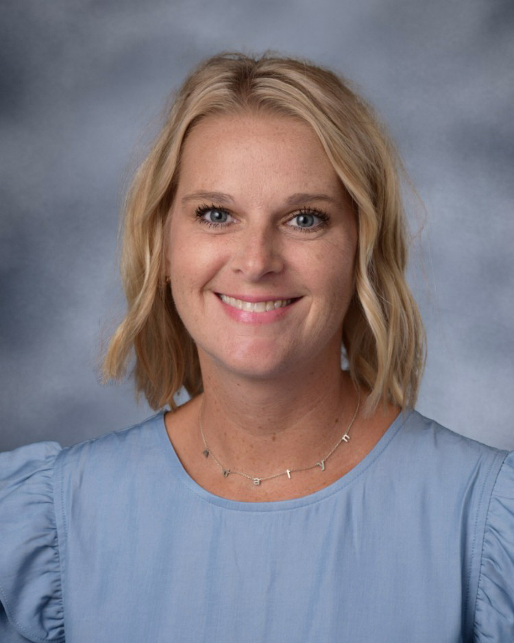 A portrait of a smiling woman with shoulder-length blonde hair and a blue blouse against a mottled grey background.