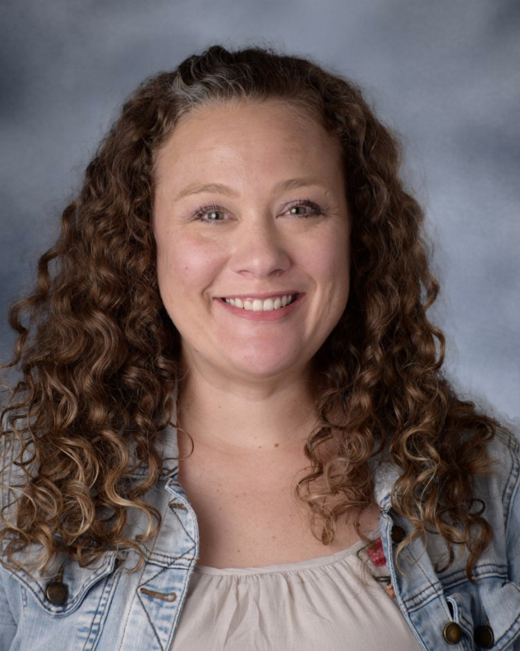 A portrait of a smiling woman with long curly brown hair wearing a denim jacket against a grey background.