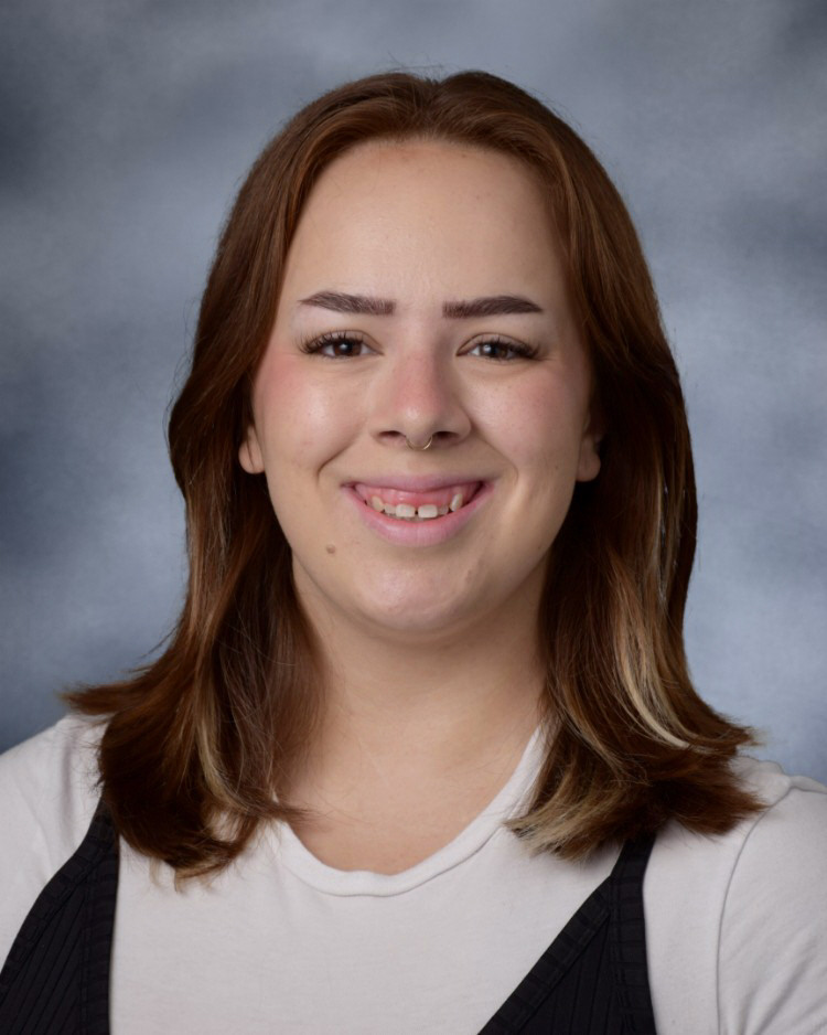 A portrait of a smiling person with reddish-brown hair and a nose ring against a grey backdrop.