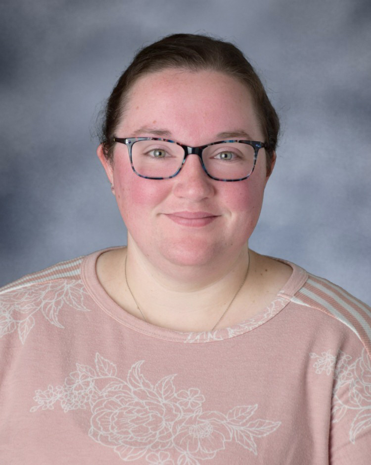 A person with brown hair and glasses wears a pink floral shirt against a grey background.
