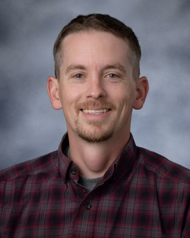 A headshot of a smiling man with short brown hair, a goatee, and a red and black plaid shirt against a grey background.
