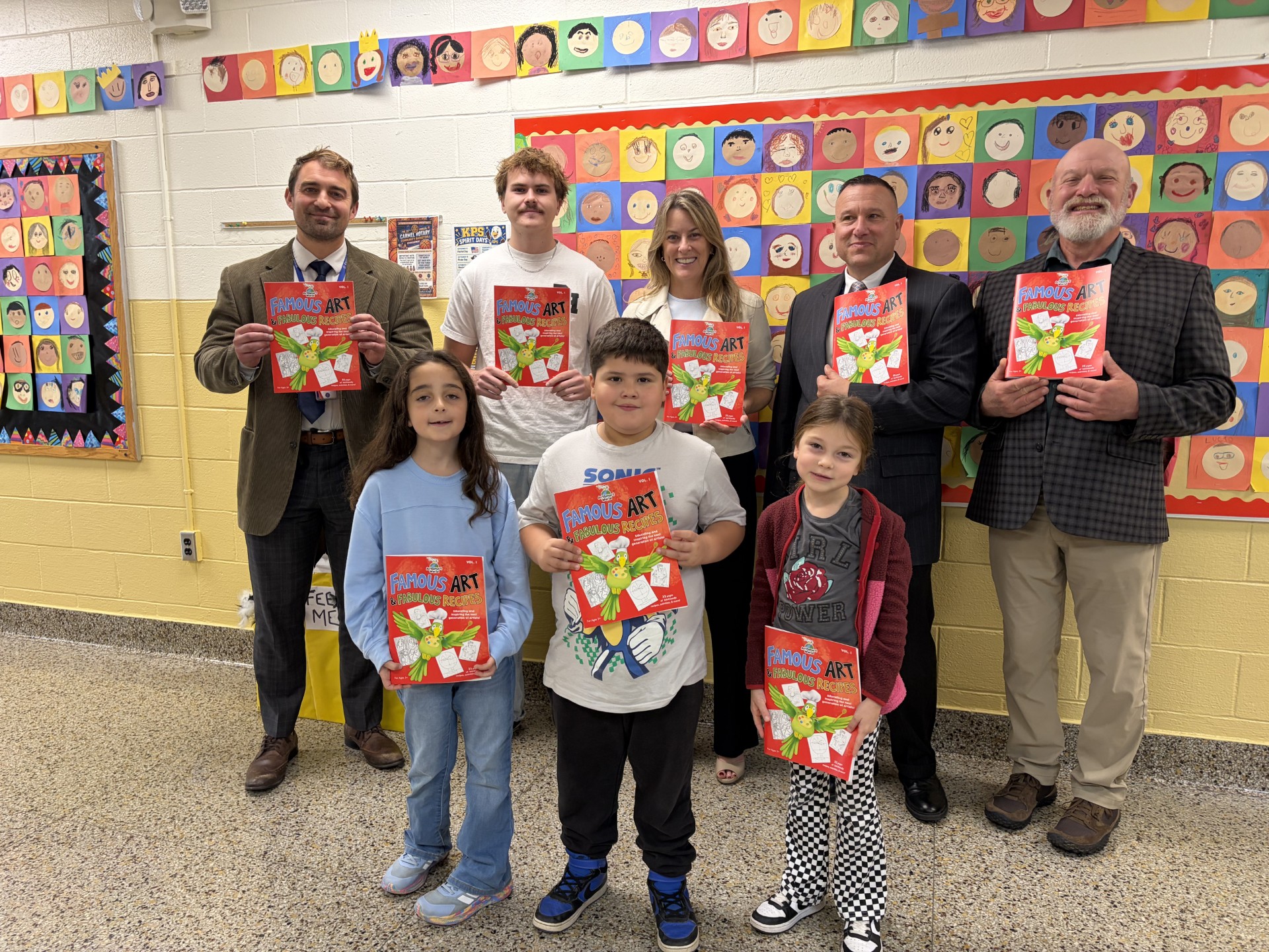 A group of adults and children proudly hold up copies of the book "Famous Art & Fabulous Recipes" in a school hallway decorated with children's artwork.