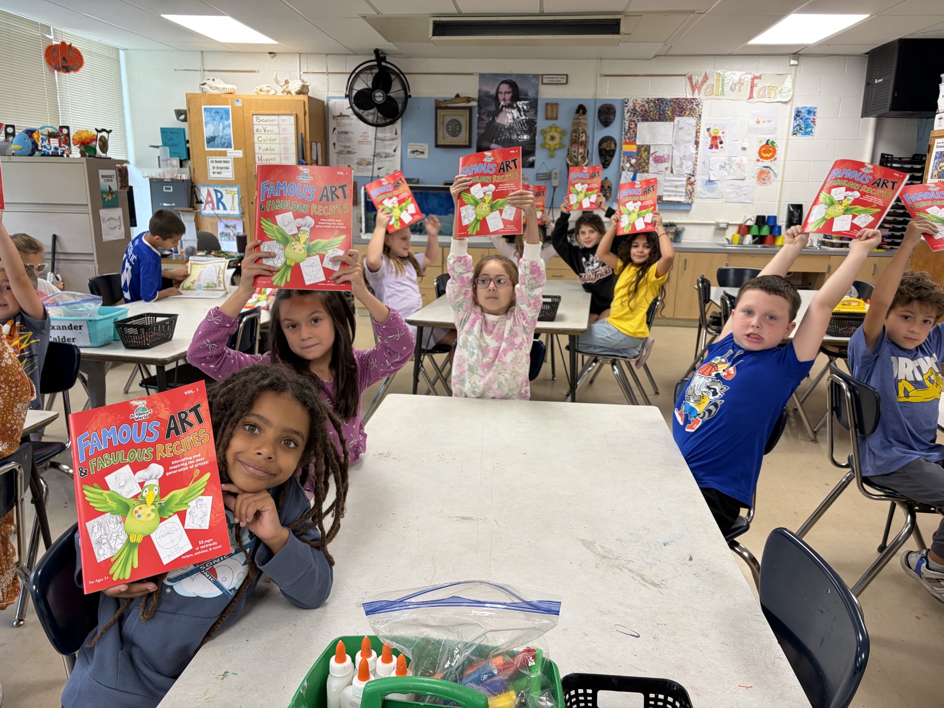 A group of elementary school students eagerly hold up copies of a book titled "Famous Art & Fabulous Recipes" in an art classroom.
