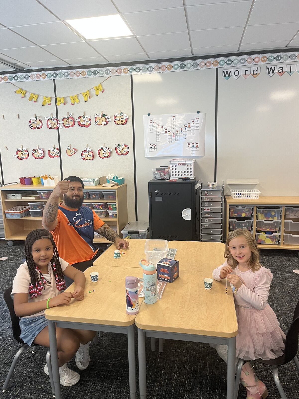 A teacher and two students sit at wooden desks in a bright classroom, working on crafts together.