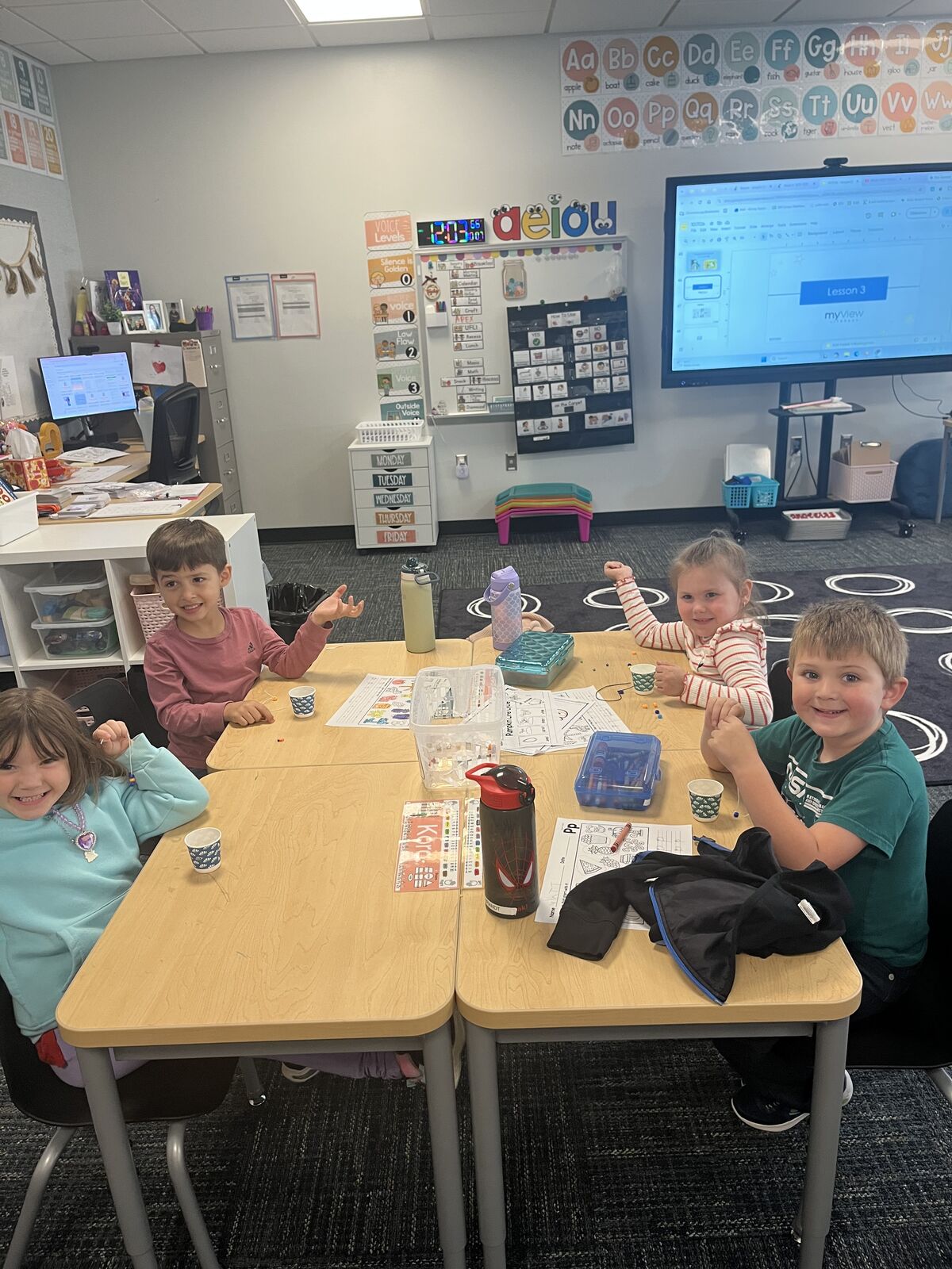 Four children sit at connected wooden desks in a bright classroom, smiling up toward the camera with school supplies spread on their tables.