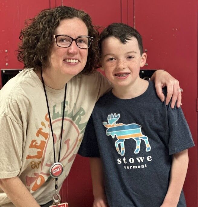 A woman with curly hair and glasses smiles next to a young boy with braces, both standing in front of red lockers.