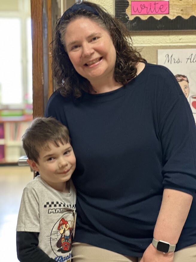 A smiling woman and a young boy stand together, with the boy in front of her.
