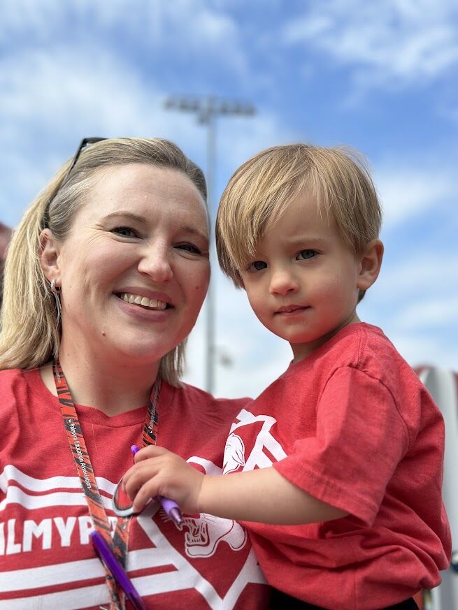 A smiling woman holds a young child, both wearing red t-shirts, outdoors under a blue sky.