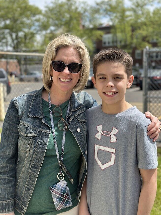 A smiling woman in a denim jacket and a young boy in a grey t-shirt stand together outdoors.