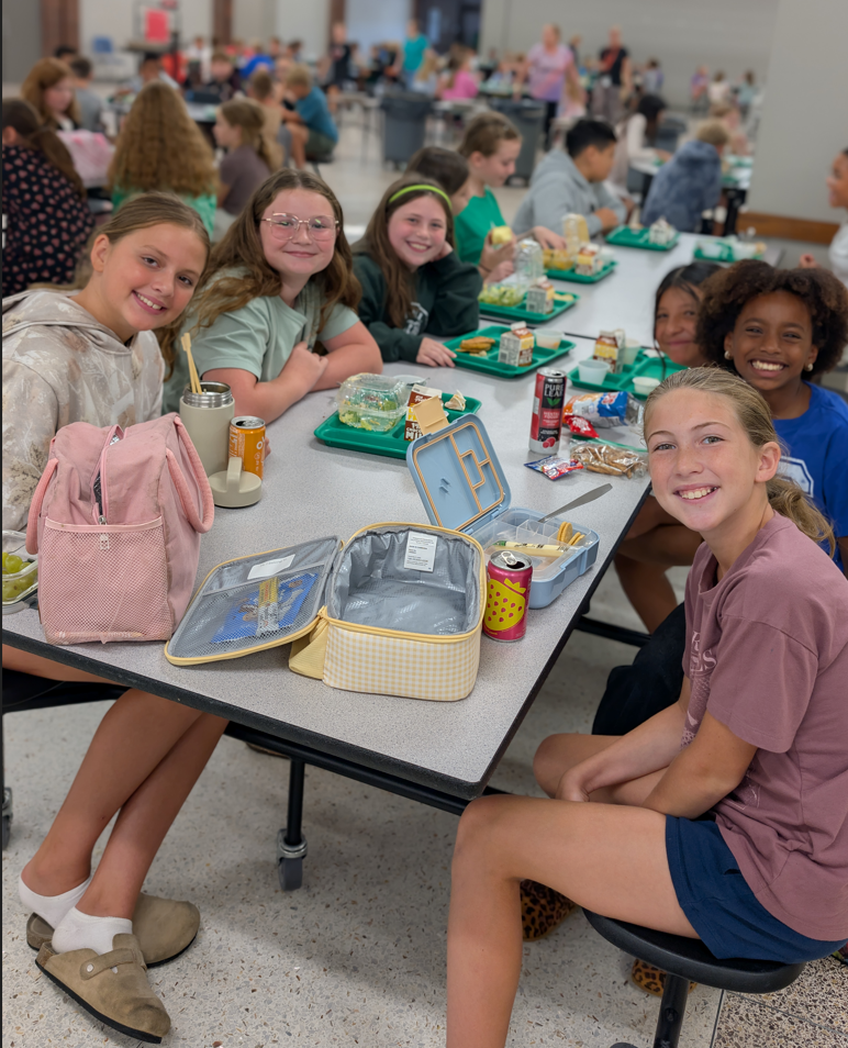 A group of smiling girls sit at a school cafeteria table with their lunches.