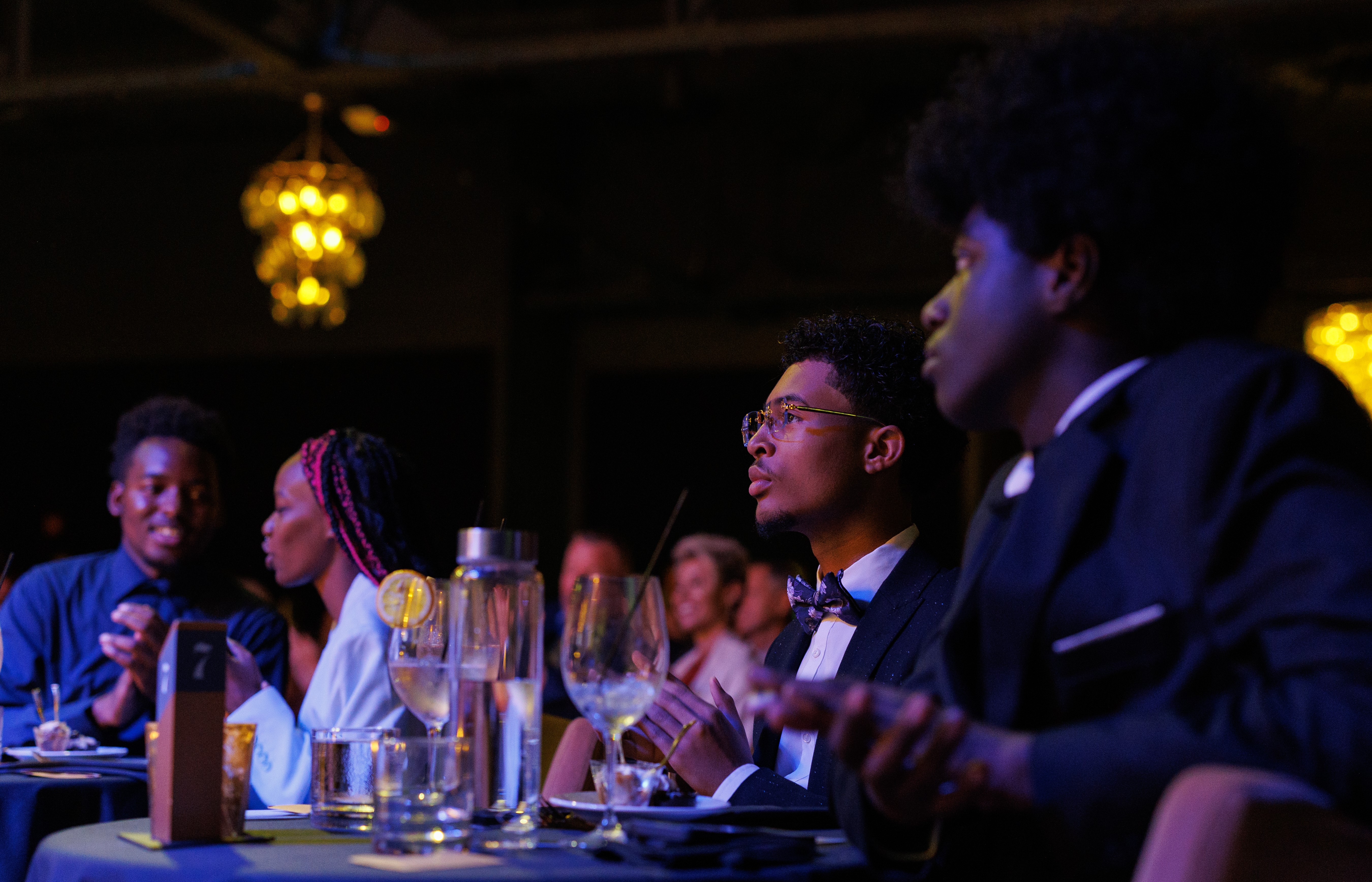 Young men and women dressed formally sit at a table with drinks and food, looking towards an unseen event.